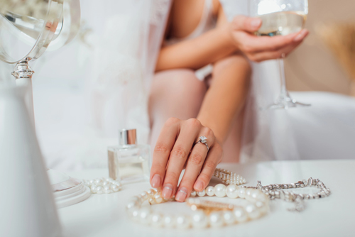 A cropped, close-up shot of a bride picking up a pearl necklace off a vanity table, with a diamond bracelet and perfume next to it.